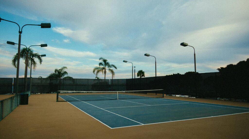 An empty outdoor tennis court surrounded by palm trees and light posts under a partly cloudy sky.